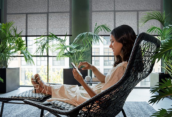 Eine Frau sitzt im Wohnzimmer auf einem Liegestuhl und guckt auf ihr Handy. Im Hintergrund sieht man eine große Fensterfront, wo die Rollos befestigt sind. Davor grüne Pflanzen.